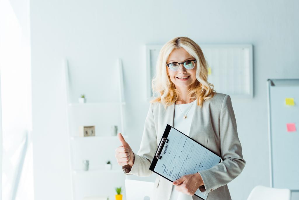 cheerful blonde woman in glasses showing thumb up while holding clipboard  - Photo, Image