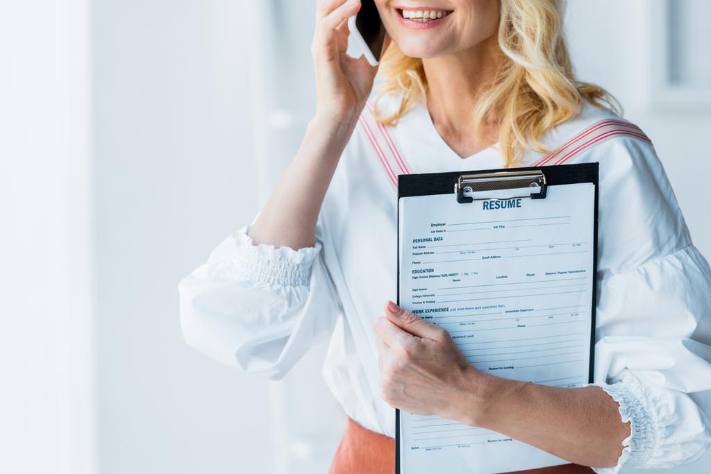 abgeschnittene Ansicht einer fröhlichen blonden Frau, die auf dem Smartphone spricht und Klemmbrett mit Lebenslauf-Briefen hält  - Foto, Bild