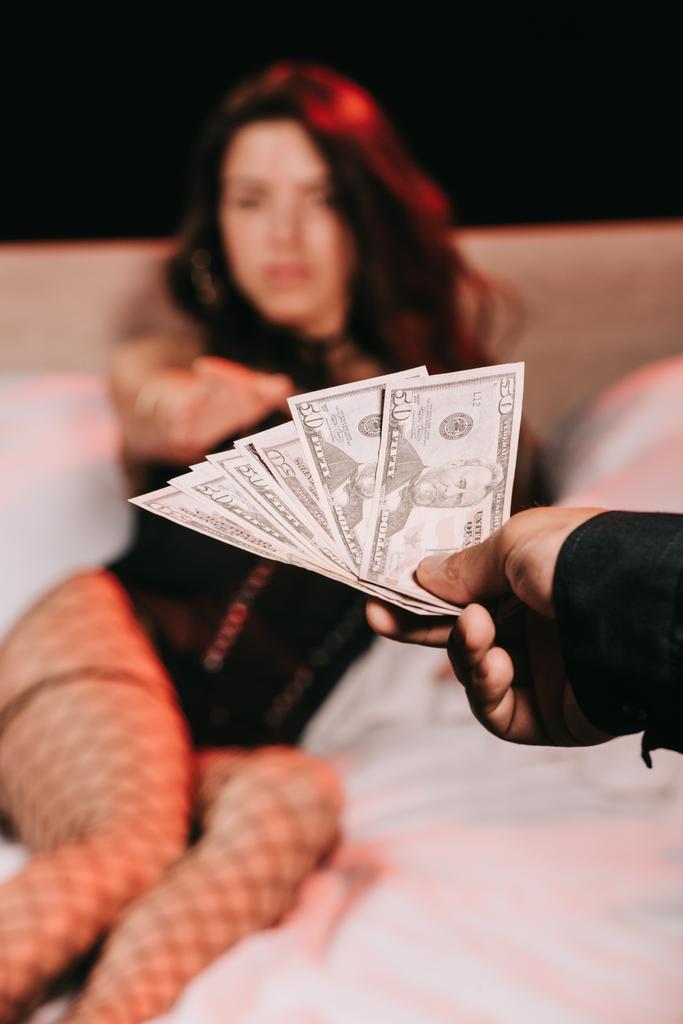 selective focus of man giving money to sexy woman in corset on bed isolated on black  - Photo, Image
