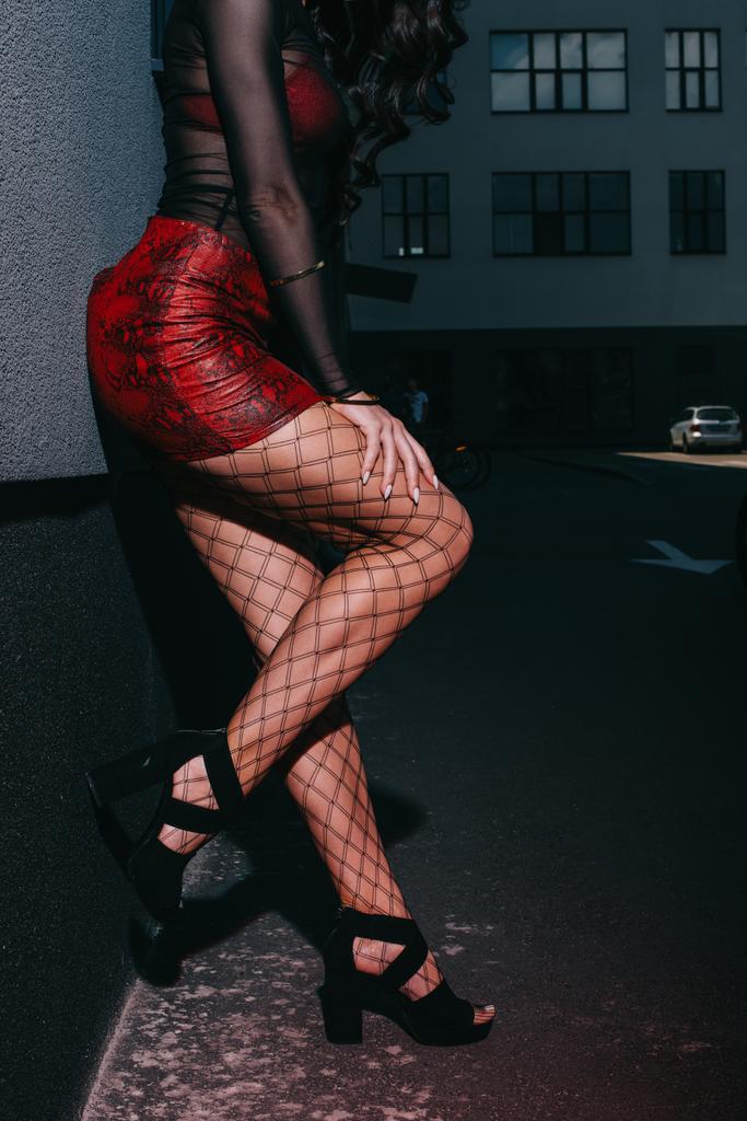 vista cortada de prostituta em saia vermelha e malha meias de pé perto da parede
  - Foto, Imagem