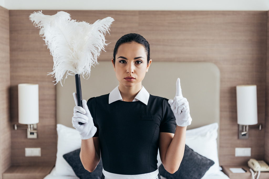 Widok z przodu poważne pokojówka w białych rękawiczkach trzyma Duster i pokazując pomysł znak w pokoju hotelowym - Zdjęcie, obraz
