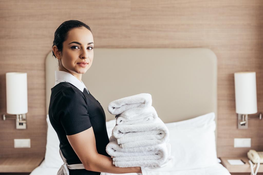 uśmiechnięta pokojówka trzyma stos złożonych ręczników w pobliżu łóżka i patrząc na kamerę w pokoju hotelowym - Zdjęcie, obraz
