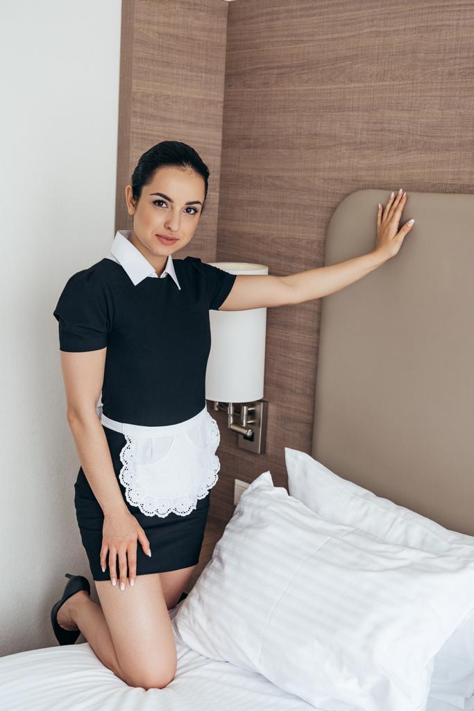 lachende meid in witte schort staande in de buurt van bed en kijken naar camera in hotelkamer - Foto, afbeelding