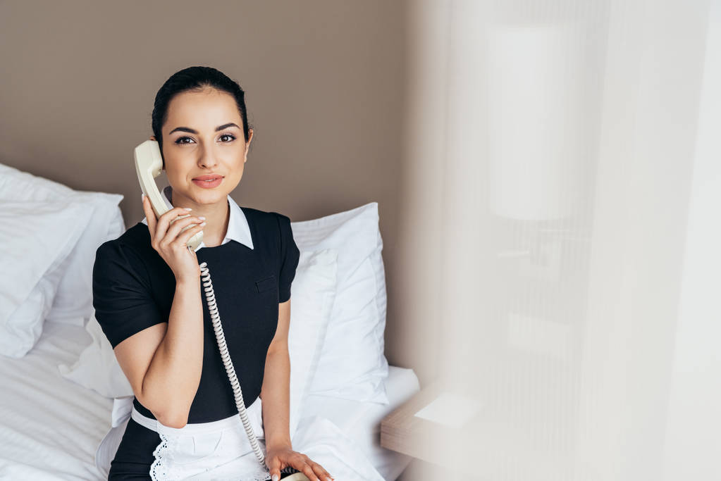 beyaz önlük güzel hizmetçi yatakta oturan ve otel odasında telefonla konuşurken - Fotoğraf, Görsel