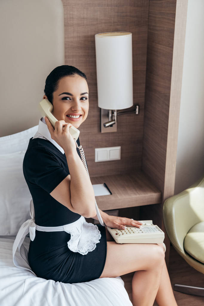 glimlachend mooi meid in witte schort zittend op bed en praten over telefoon in hotelkamer - Foto, afbeelding