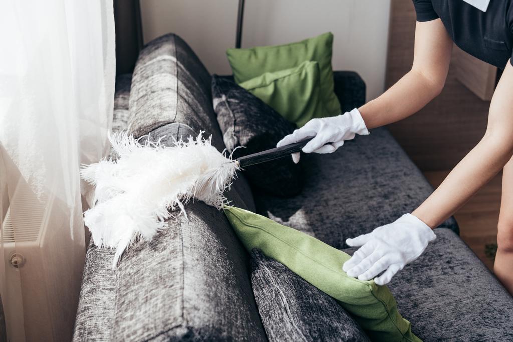 částečný pohled na služebnou v bílých rukavicích čištění pohovky s dusterem v hotelovém pokoji - Fotografie, Obrázek