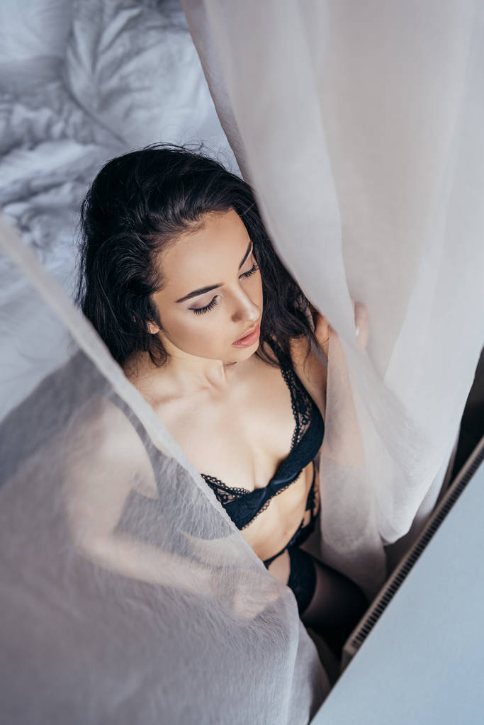 stropní pohled na sexy mladou ženu v černé podprseně blízko bílé opony - Fotografie, Obrázek