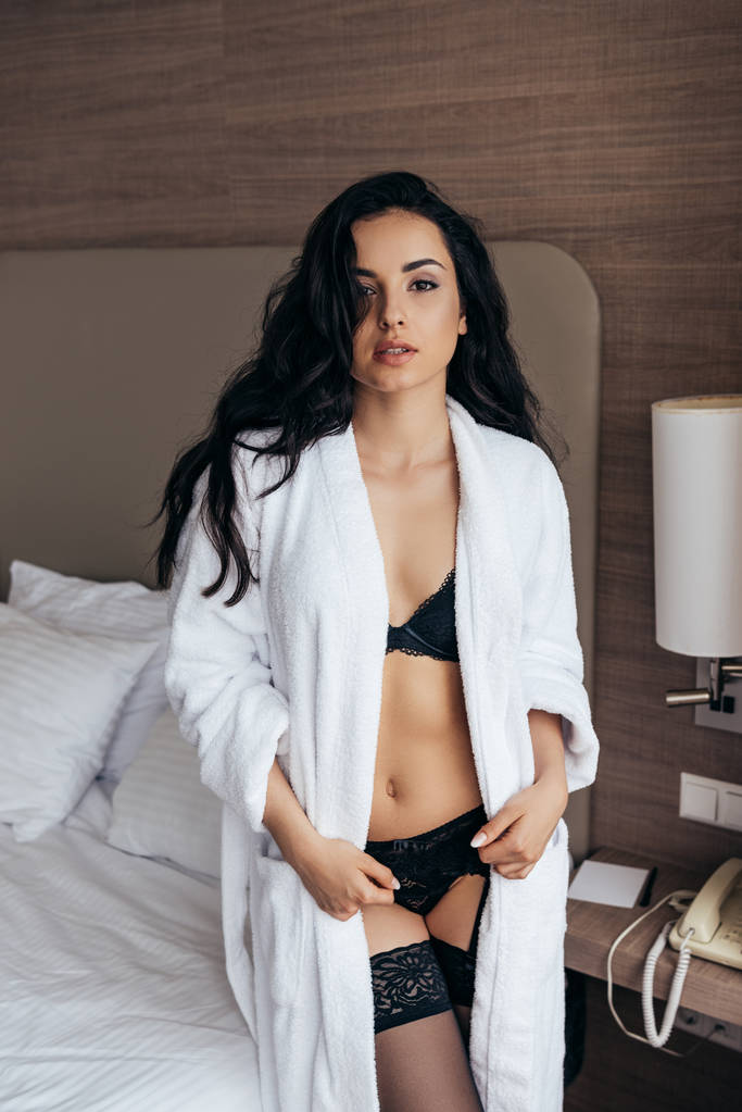sexy bruna giovane donna in lingerie e accappatoio bianco guardando la fotocamera in camera da letto
 - Foto, immagini