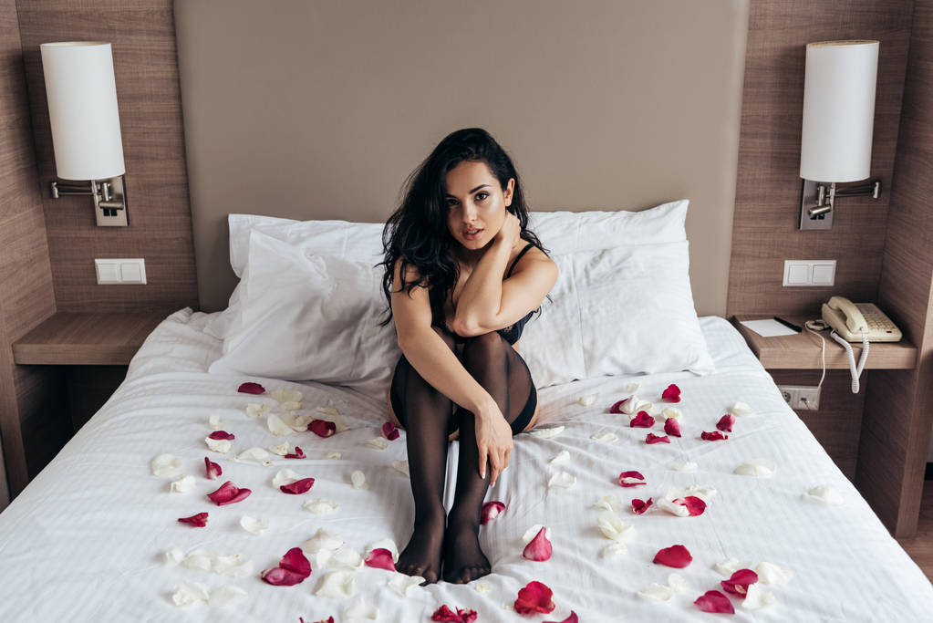 寝室でバラの花びらとベッドに座って黒いストッキングのセクシーなブルネットの女の子 - 写真・画像
