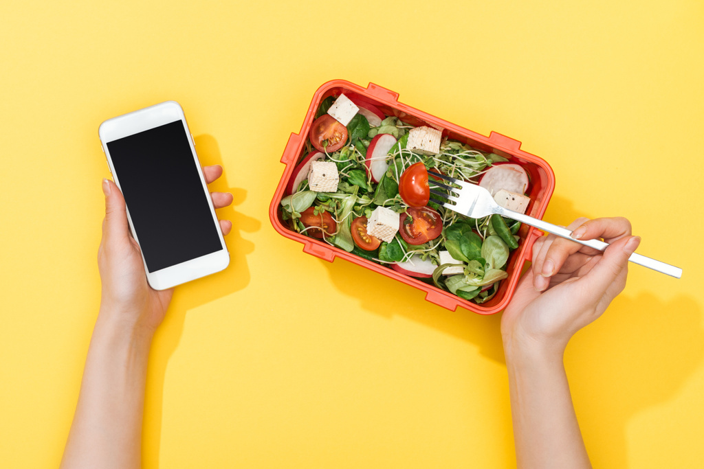 обрезанный вид женщины с вилкой и обеденная коробка с салатом с помощью смартфона
 - Фото, изображение