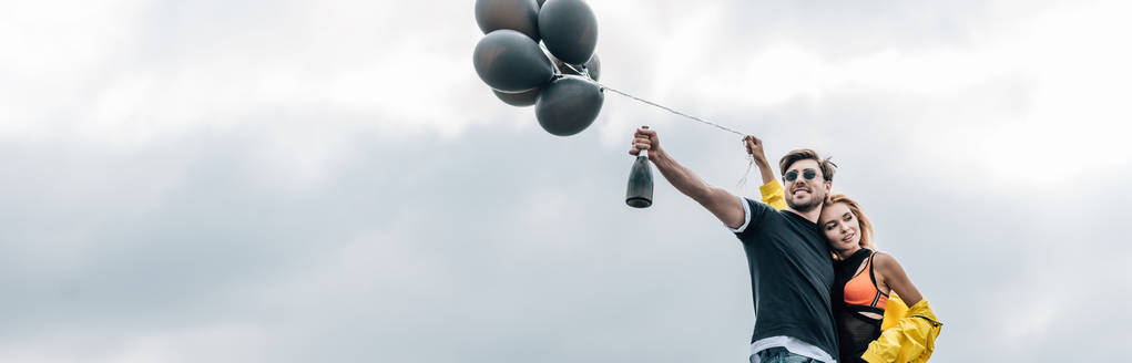 panoramische shot van man Holding fles en aantrekkelijke vrouw houden ballonnen  - Foto, afbeelding