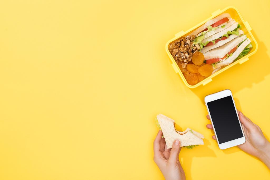 oříznutý pohled na ženu držící sendvič a smartphone v okolí krabice s jídlem - Fotografie, Obrázek