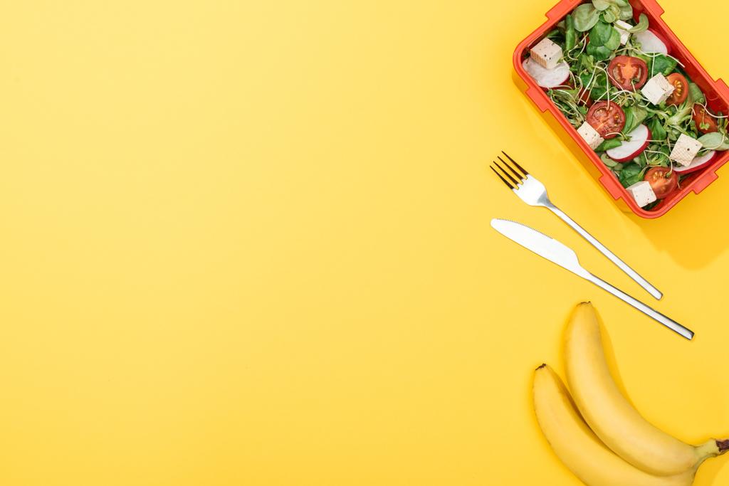 サラダ付き弁当箱の近くにバナナ、フォーク、ナイフのトップビュー - 写真・画像
