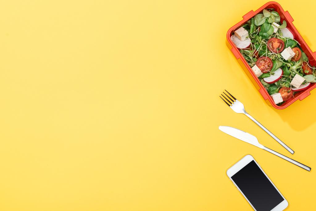 вид зверху обідньої коробки з салатом біля виделки, ножем і смартфоном
 - Фото, зображення