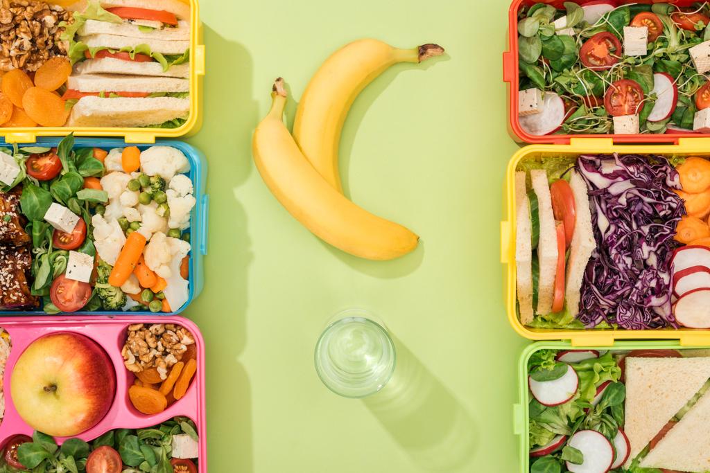 вид на обеденные коробки с едой возле стакана воды и бананов
 - Фото, изображение