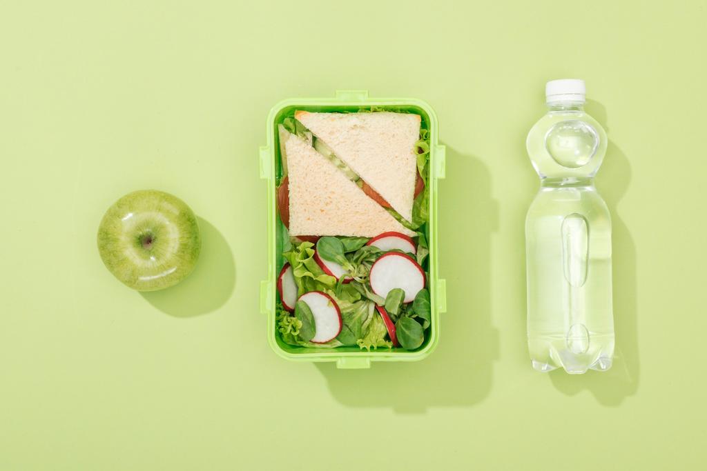 Κορυφαία θέα στο κουτί του μεσημεριανού γεύματος με σαλάτα και σάντουιτς κοντά σε μπουκάλι με νερό και πράσινο μήλο - Φωτογραφία, εικόνα