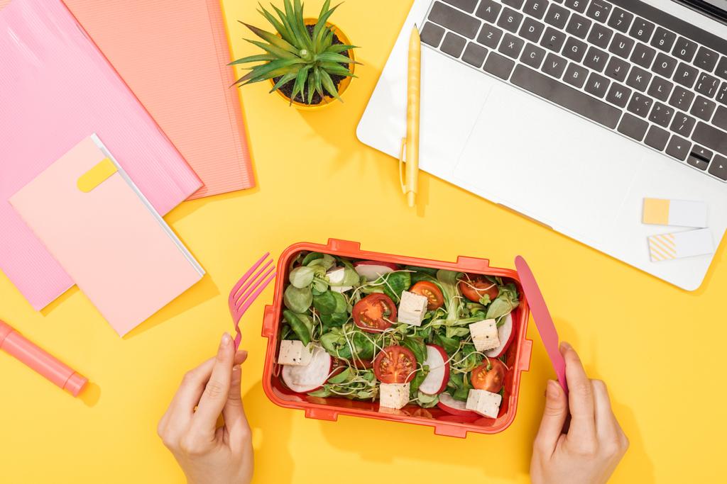 vue recadrée de la femme tenant fourchette sur la boîte à lunch avec de la nourriture près d'un ordinateur portable et des fournitures de bureau
 - Photo, image