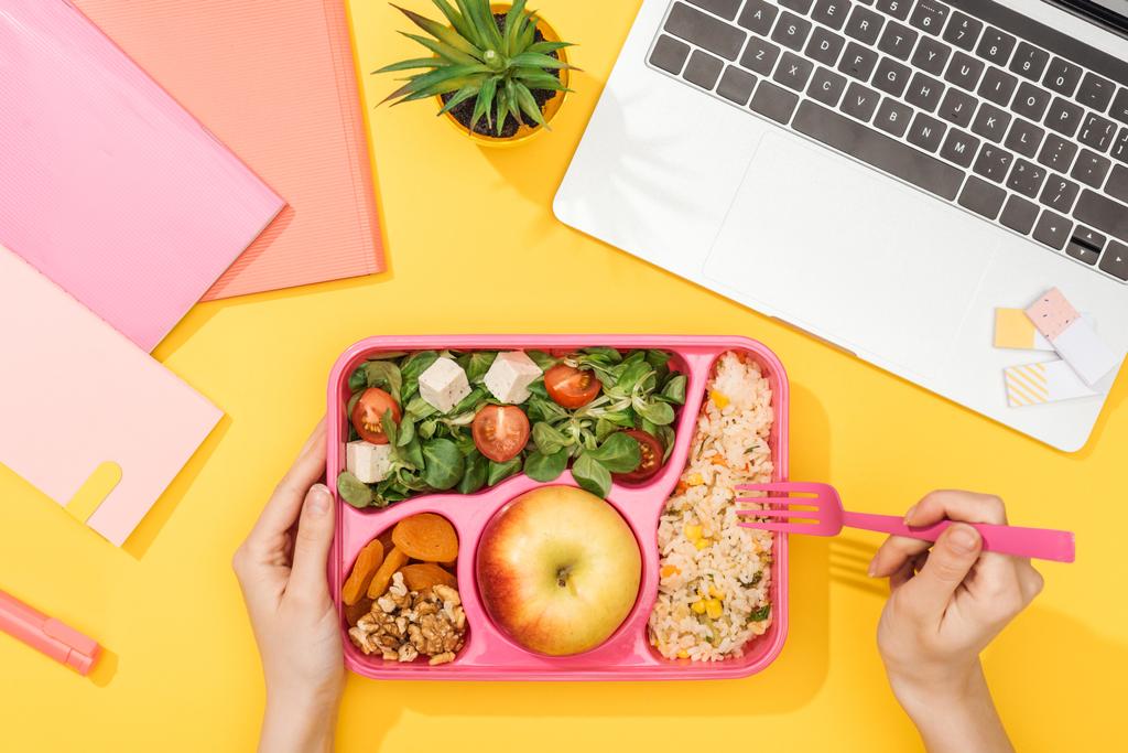 Ausgeschnittene Ansicht einer Frau mit Gabel über Lunchbox mit Lebensmitteln in der Nähe von Laptop und Bürobedarf - Foto, Bild