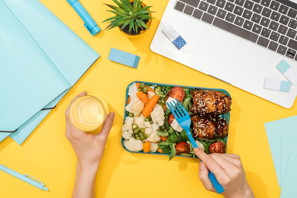 vágott kilátás nő gazdaság ebéd doboz élelmiszer-és pohár víz közelében laptop és irodai kellékek - Fotó, kép