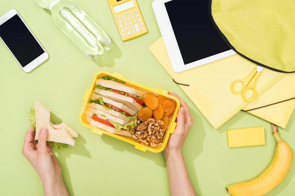 levágott kilátás nő kezében szendvics, műanyag edények több mint ebéd doboz élelmiszer közelében hátizsák, digitális tabletta, üveg víz és írószer - Fotó, kép