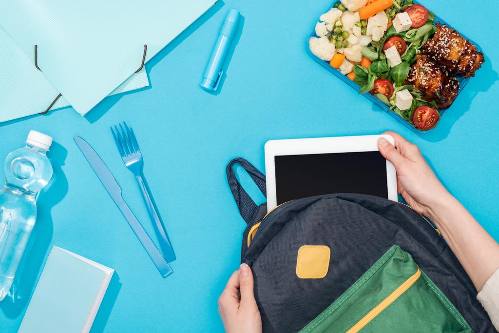 bijgesneden beeld van de vrouw verpakking digitale Tablet in rugzak in de buurt van lunch box, briefpapier en fles water - Foto, afbeelding