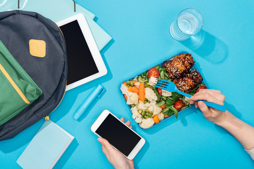 cropped view of woman hands with fork and smartphone near lunch box with food, backpack, digital tablet, glass of water and stationery - Photo, Image