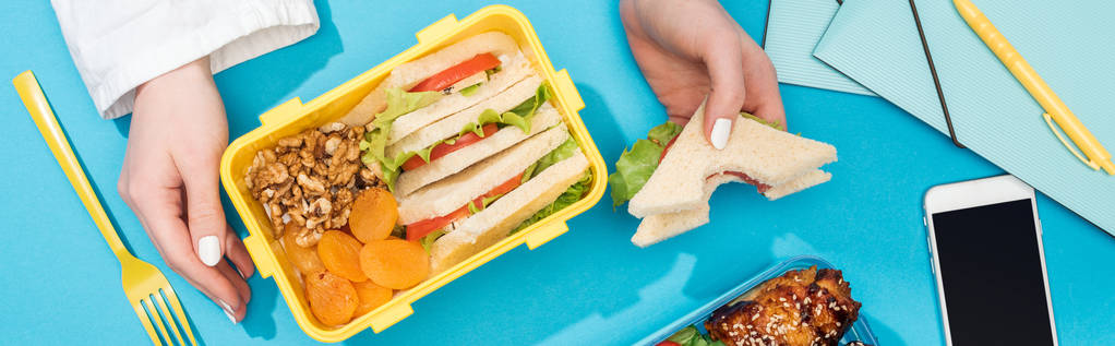 Ausgeschnittene Ansicht einer Frau mit Lunchbox und Sandwich in der Hand in der Nähe von Smartphone und Bürobedarf - Foto, Bild