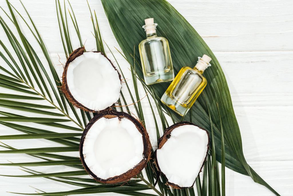Top uitzicht van kokosolie in flessen op groene Palm bladeren op wit houten oppervlak - Foto, afbeelding
