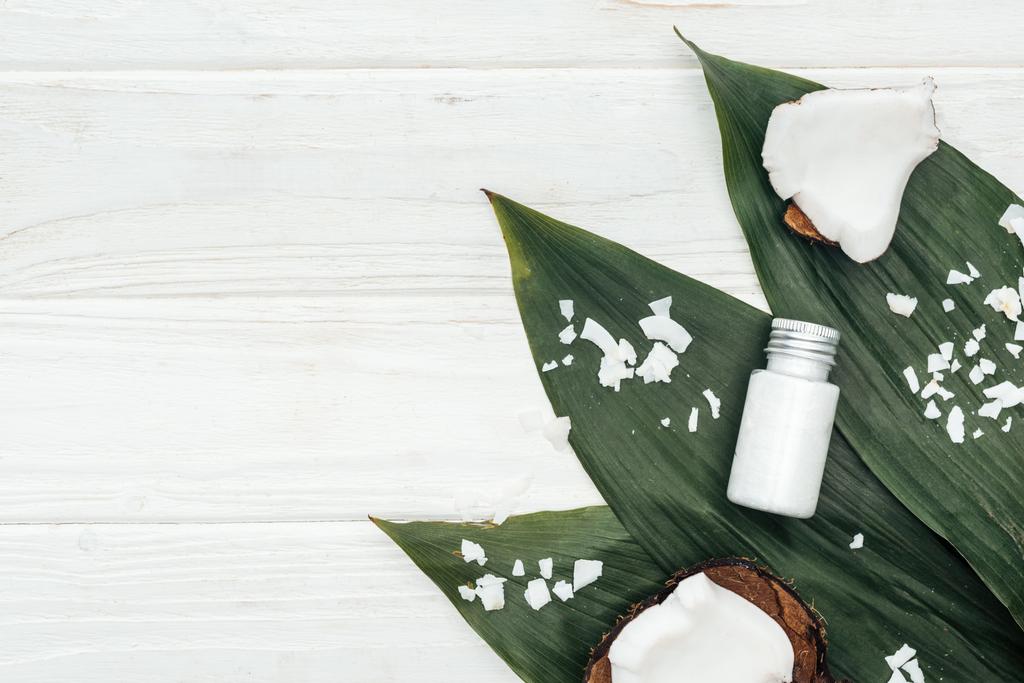 pohled na produkt z kokosových krás v láhvi na zelených palmových listech s kokosovými vločkami na bílém dřevěném povrchu s prostorem pro kopírování - Fotografie, Obrázek