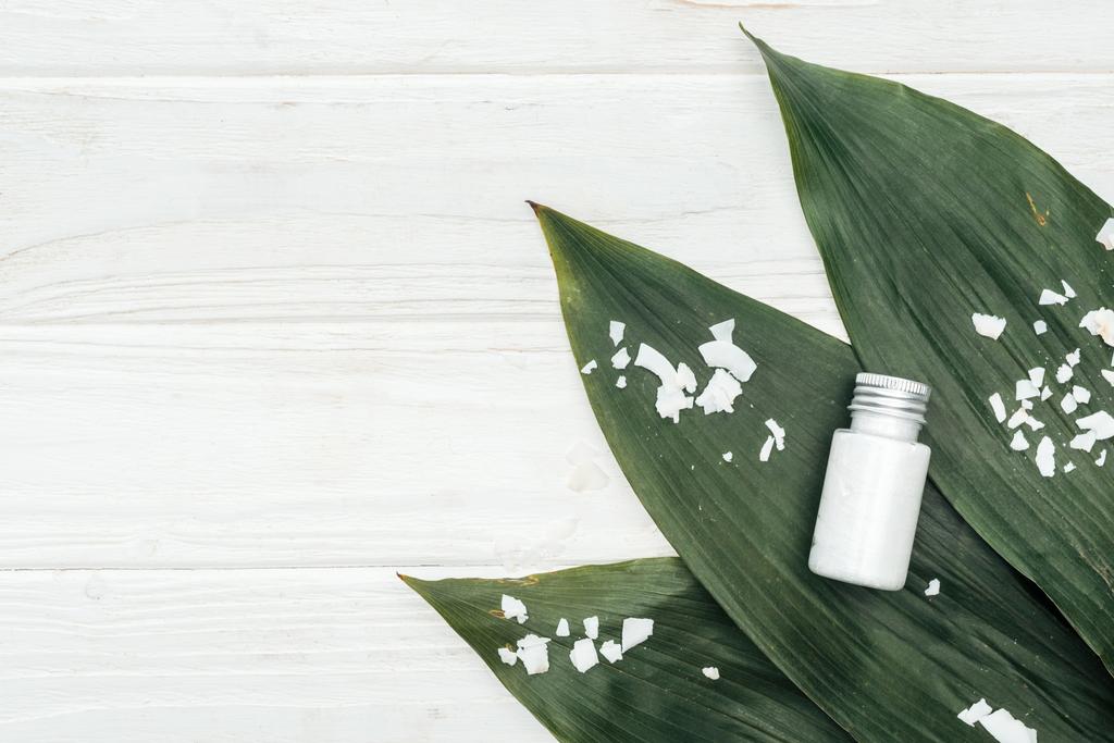 Κορυφαία θέα του προϊόντος ομορφιάς καρύδας σε μπουκάλι σε πράσινα φύλλα παλάμης με νιφάδες καρύδας σε λευκή ξύλινη επιφάνεια - Φωτογραφία, εικόνα