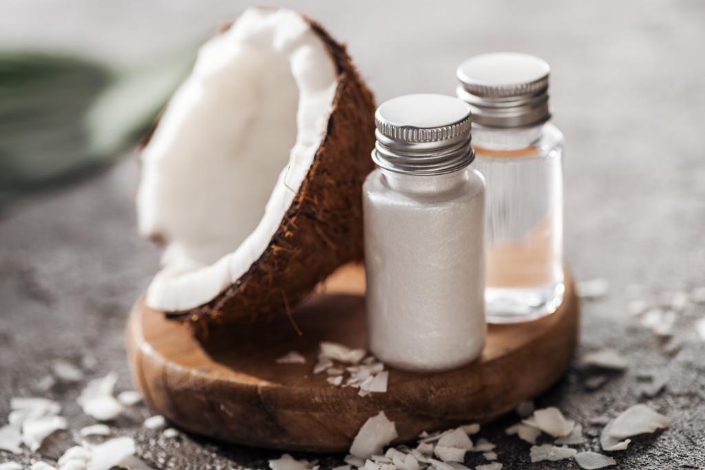 selektivní zaměření lahví s výrobky z kokosových krás na dřevěné desce blízko kokosového ořechu napůl na šedém texturově pozadí - Fotografie, Obrázek