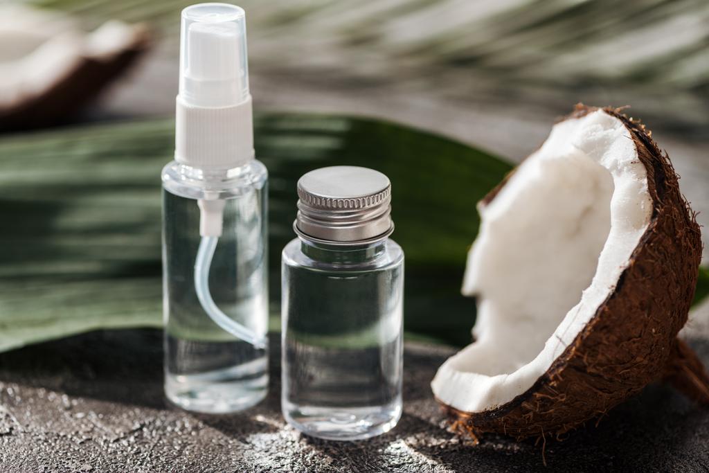 Kookosvettä sisältävien pullojen valikoiva fokus kookospähkinän puolikkaan lähellä ja palmunlehti harmaalla kuvioidulla pohjalla
 - Valokuva, kuva