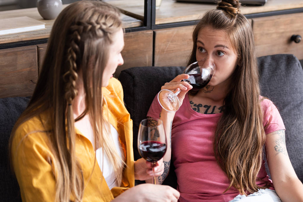 twee lesbiennes houden wijn glazen en kijken naar elkaar terwijl zittend op de Bank in de woonkamer - Foto, afbeelding