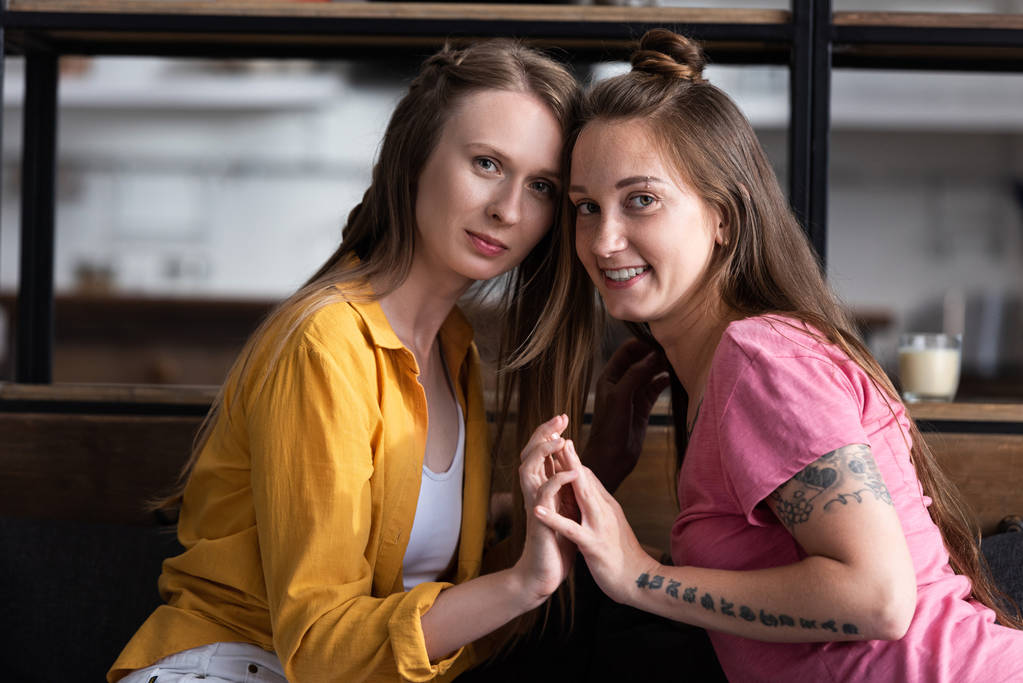 dva usmívající se lesbičky dotýkali se rukou a dívali se na kameru v obývacím pokoji - Fotografie, Obrázek