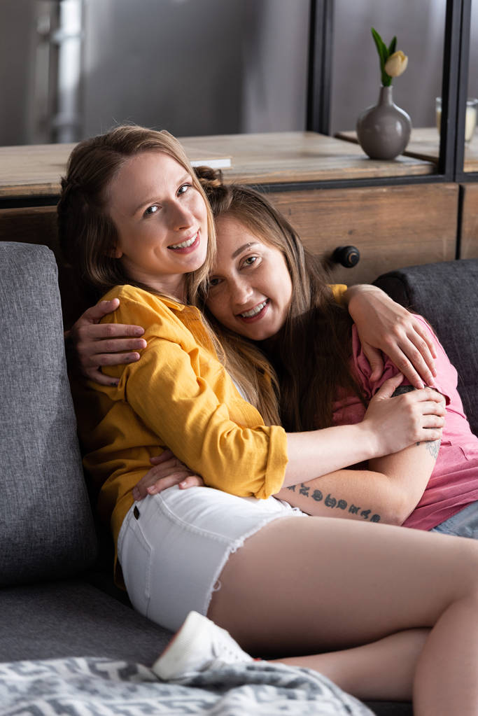 zwei hübsche Lesben umarmen sich, während sie auf dem Sofa im Wohnzimmer sitzen - Foto, Bild
