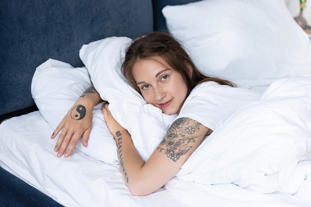 привлекательная девушка с татуировками, лежащая в постели и смотрящая на камеру в спальне
 - Фото, изображение