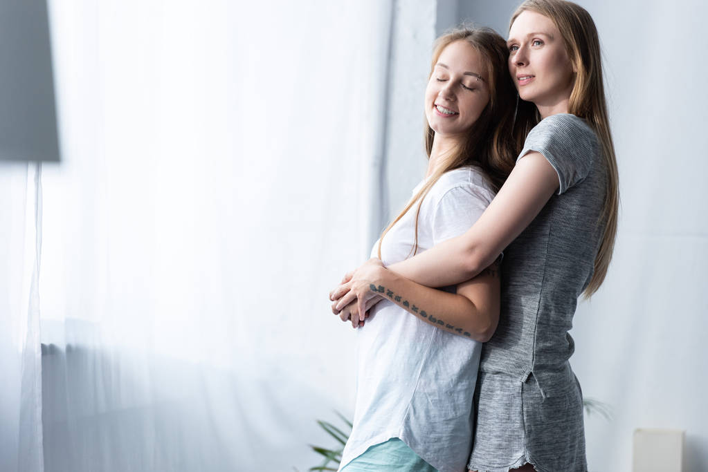 dos lesbianas sonrientes en camisetas abrazándose en el dormitorio
 - Foto, Imagen