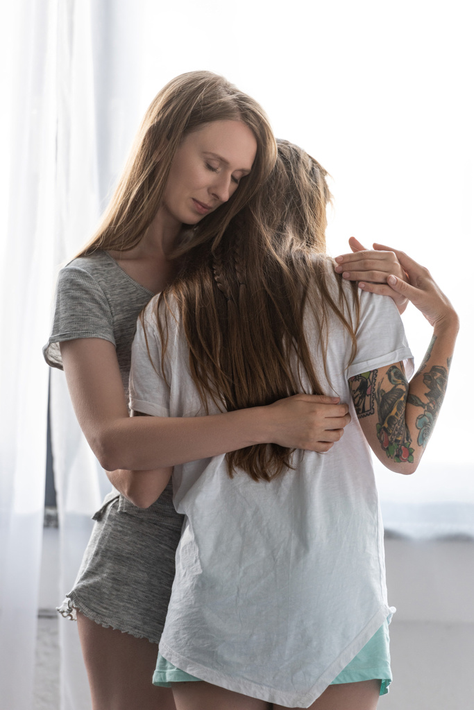 twee glimlachende lesbiennes in t-shirts omarmen in slaapkamer - Foto, afbeelding