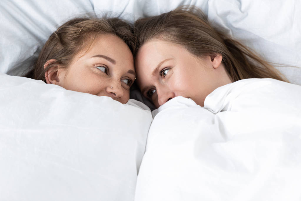 két leszbikusok alatt fekvő fehér takaró és nézi egymást a hálószobában - Fotó, kép