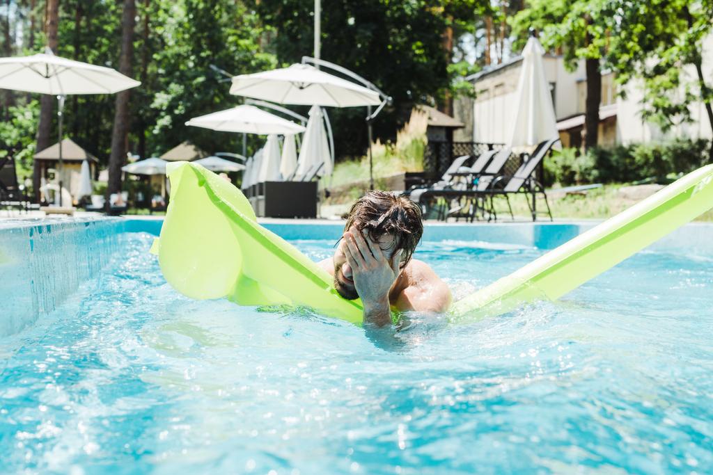 bebaarde man zwemmen in zwembad met groene opblaasbare ring en bedekken gezicht met handen  - Foto, afbeelding