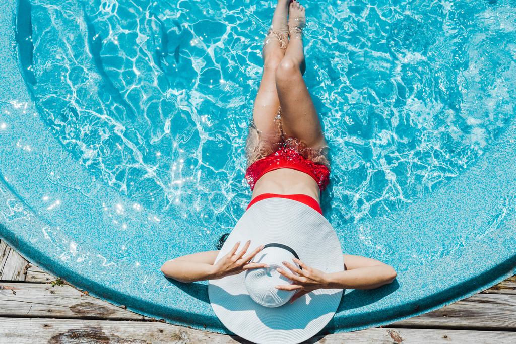 麦わら帽子に触れ、青い水の中で赤い水着に座っている女性のトップビュー  - 写真・画像