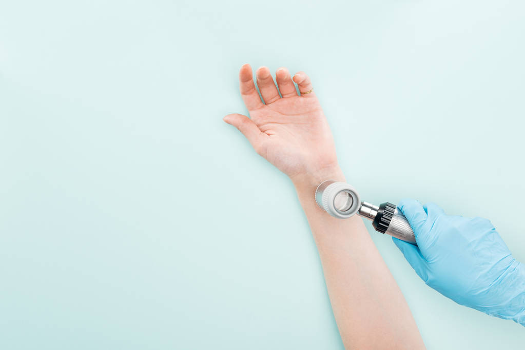 bijgesneden beeld van arts onderzoeken hand van vrouw met dermatoscoop geïsoleerd op blauw - Foto, afbeelding