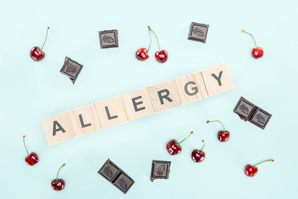 Ansicht von Holzblöcken mit Allergie-Schriftzug in der Nähe von Kirschen und Schokolade isoliert auf blau - Foto, Bild