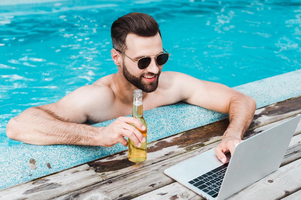 vrolijke bebaarde man in zonnebril met laptop terwijl het houden van fles met bier  - Foto, afbeelding