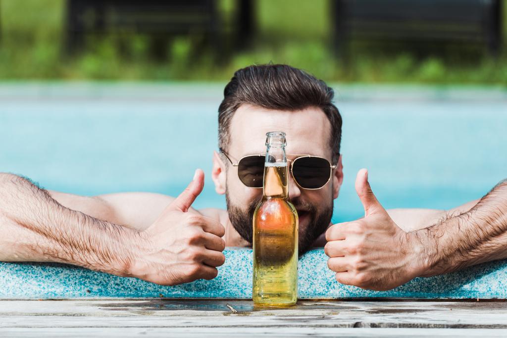 Bärtiger Mann mit Sonnenbrille zeigt Daumen hoch neben Flasche mit Bier  - Foto, Bild
