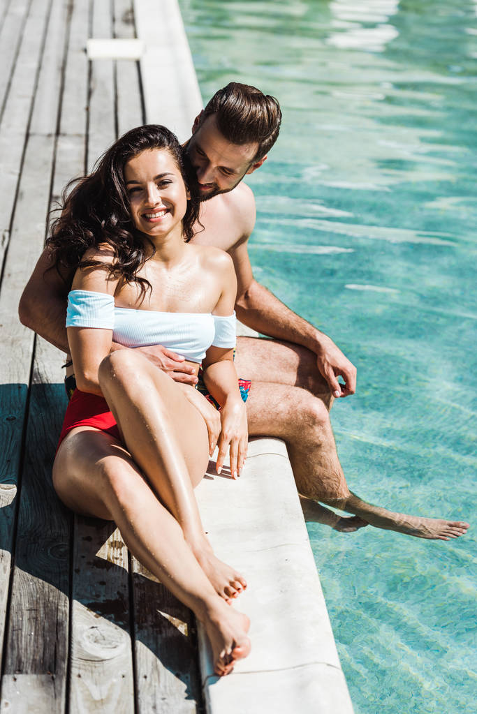 šťastný muž, který sedí s veselou ženou na dřevěných palubách blízko bazénu  - Fotografie, Obrázek