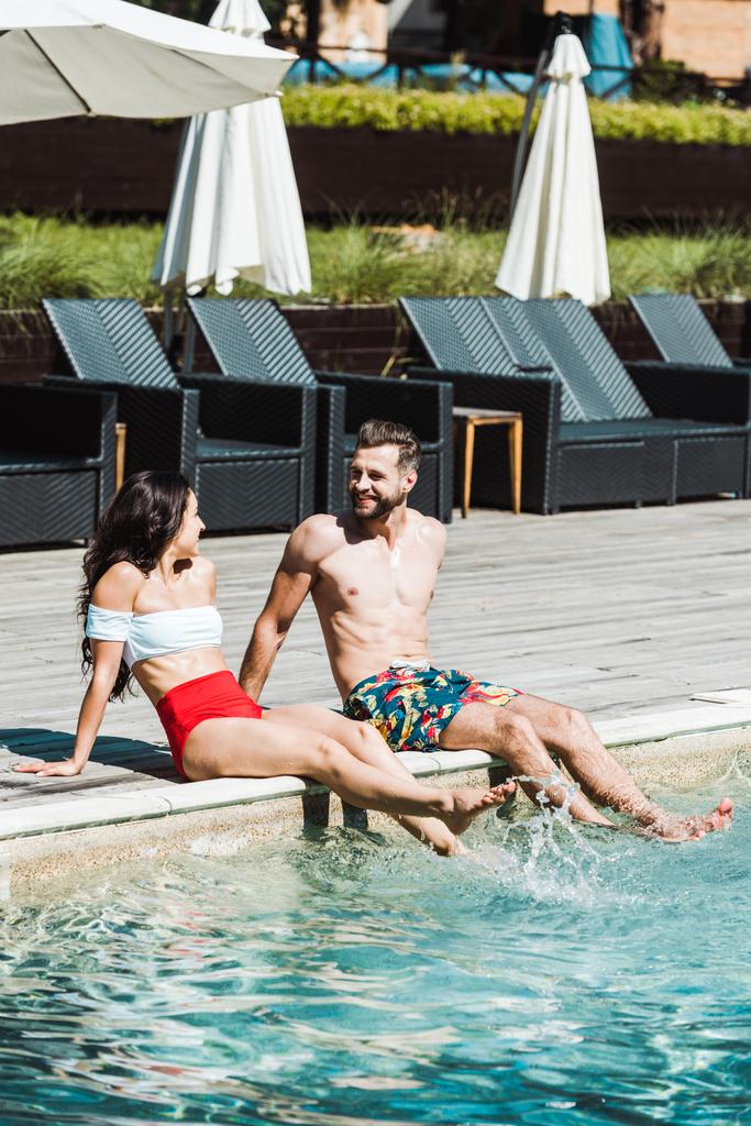 hombre alegre mirando a la mujer mientras está sentado en cubiertas de madera cerca de la piscina
  - Foto, imagen