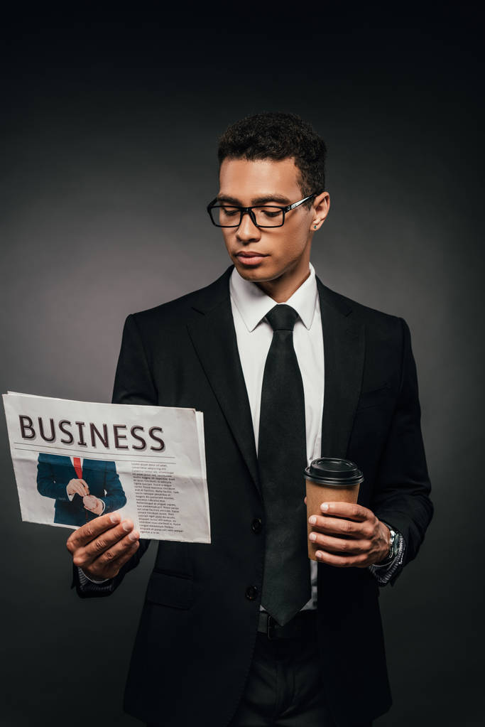 красивий афроамериканець бізнесмен в окулярах та костюмі проведення кави піти і читати газету на темному фоні - Фото, зображення