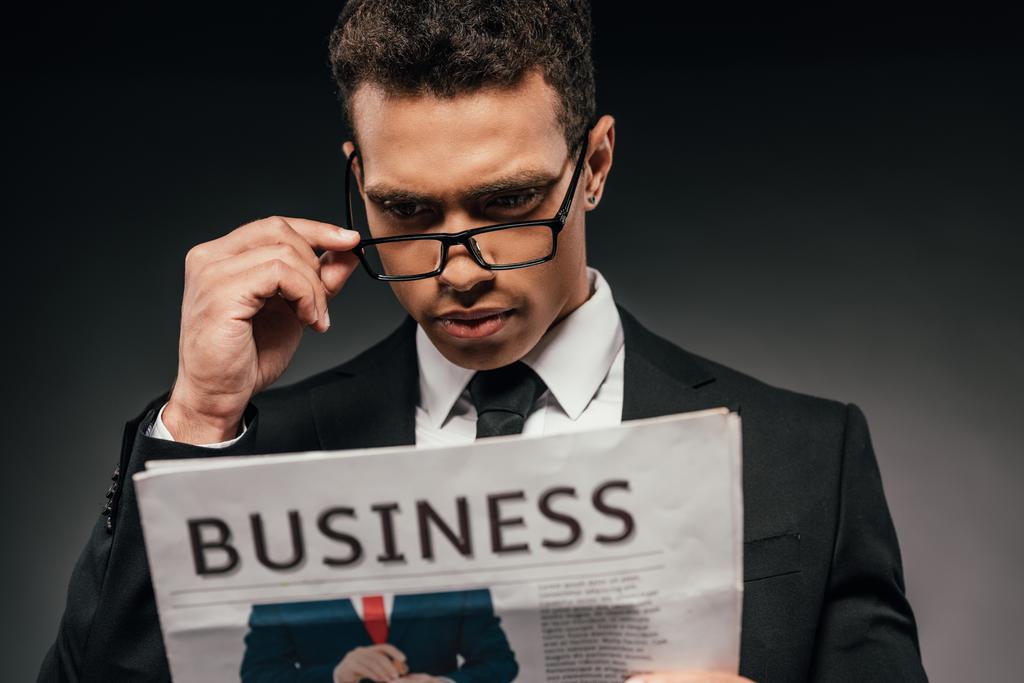 Африканский американский бизнесмен в очках и костюме читает деловую газету на тёмном фоне
 - Фото, изображение
