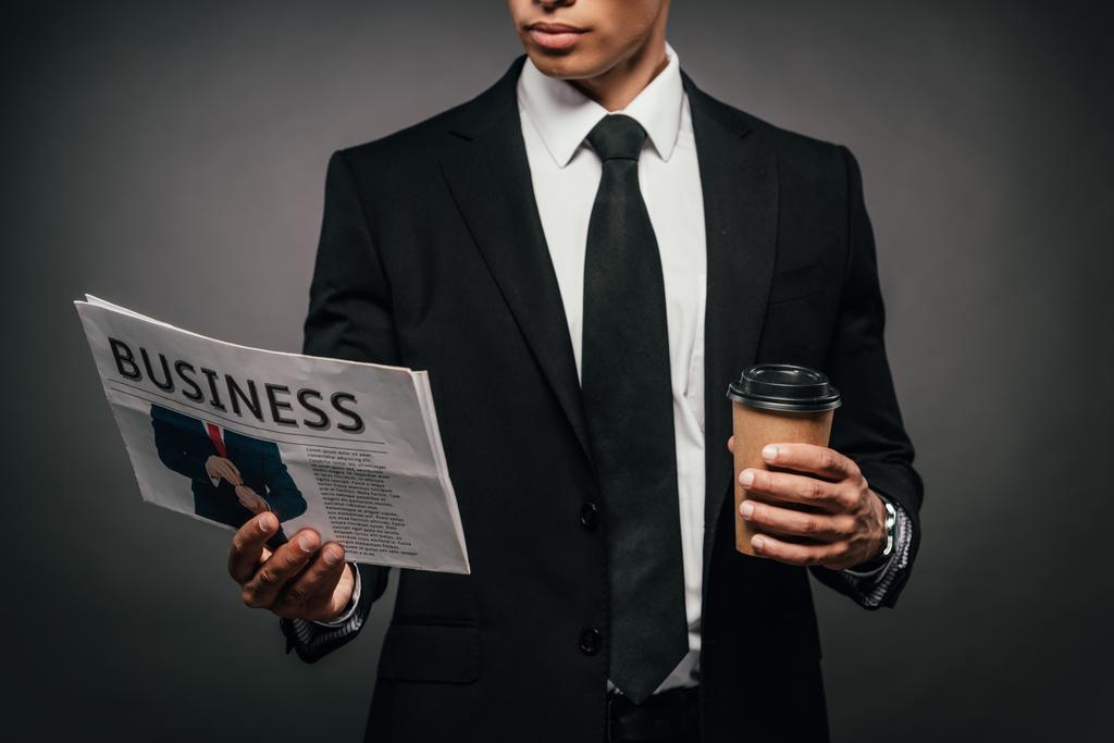 visão parcial de belo empresário afro-americano em terno ler jornal de negócios e segurando café para ir em fundo escuro
 - Foto, Imagem